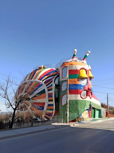 Photography. A color photo of a five-storey house that looks like a rainbow snail with a shell. Painted in shades of red, orange, green, blue and brown, the house has "no straight walls, corners or edges" and yet is a "normal" house. With two tentacles on the head and curved windows on the sides, the snail was designed in such a way that all parts of the house appear to be part of the snail's body. The door of the snail shell is painted as the snail's mouth. The normal chimney has been replaced by a large yellow bee on the snail's back. The bee's horns not only channel the smoke away, but also serve as a night light and lightning rod. The eyes have a purpose in the snail's shell. The air and gases of the snail's shell flow in through the eyelids and exit through the snail's large red eyes. Info: The heaters in the snail shell are disguised as frogs, ladybugs and pumpkins.The Snail House is constructed entirely with lightweight and eco-friendly material, making it truly energy efficient. 