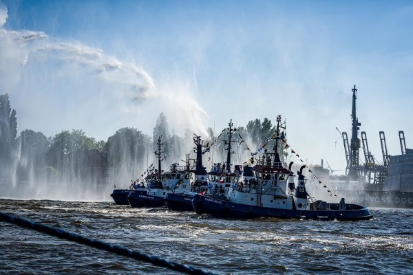 Four tugboats sitting side by side with a fireboat spraying water in the background during the annual harbour birthday in Hamburg and its traditional tugboat ballet