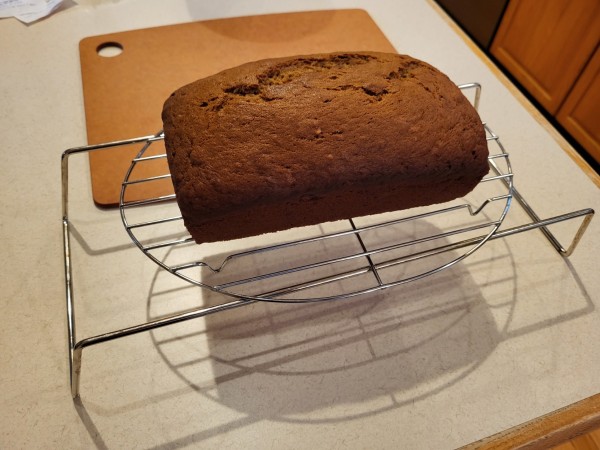 A load of dark brown banana bread rests on a wire cooling rack, next to a cutting board.