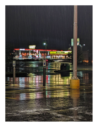 night. lit by streetlights. in the foreground an empty, wet supermarket parking lot with light pole. in the mid-distance, a waffle house with one car in front, headlights on. across the highway, a circle k store with lighted, red fuel canopy in front. rain is visible.