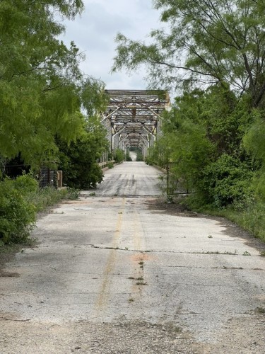 A dilapidated road leading to an old, rusted, metal truss bridge, flanked by overgrown greenery under an overcast sky.