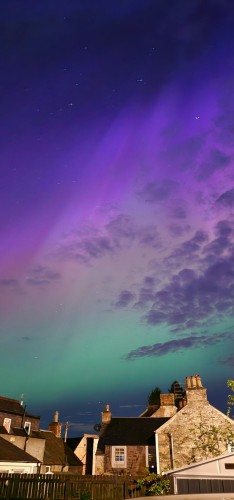 A colour photograph of an aurora in the sky above houses in a village. From turquoise at the bottom of the sky through yellow to purple at the top.