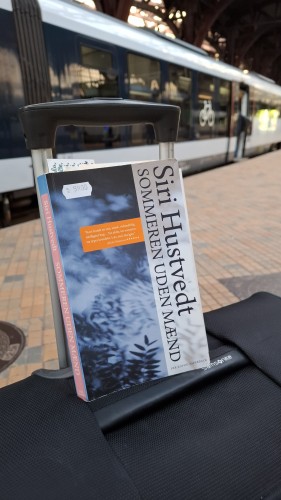 Propped up on a suitcase on the platform of the railway station, a train visible in the background: my paperback edition of Siri Hustved's novel Sommeren uden mænd. (The Summer Without Men), a bookmark sticking out. The cover is a generic dull abstract 'leafy shadows on blue' image on the left. Perked up on the right by the author name and title as a vertical panel. A 59 kr. sticker reveals this is a second-hand book, cheap by Danish book price standards. 