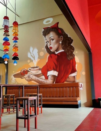 Streetartwall. On the wall of a café in a large business center is a cool mural featuring a pinup girl. The red-haired beauty is looking over her shoulder and holding a tray with a fragrant cup of coffee and a donut. She is wearing a red waitress outfit, a red bow in her hair and a small halo over her head. She has red lips and a kissy mouth. Her arm is tattooed (in the photo you can see a long row of brown leather seats, some bar stools and two colorful decorative wind chimes hanging from the ceiling).