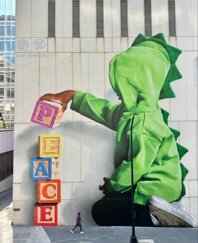 Streetartwall. A huge mural of a little boy building a tower with the word "peace" using building blocks was sprayed on the outside wall of a modern four-storey building. The little boy can be seen from the side. He is sitting in a green hootie in front of a tower of blocks with colorful letters on them. He lifts the last block with the "P" onto the top of the others.
Title: "Rebuilding Blocks"