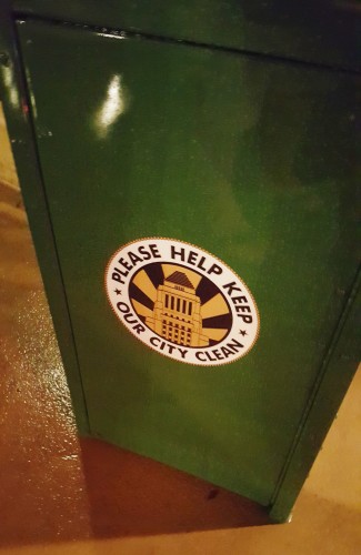 A green trash can with a sticker that reads "Please Help Keep Our City Clean" accompanied by an illustration of a building.