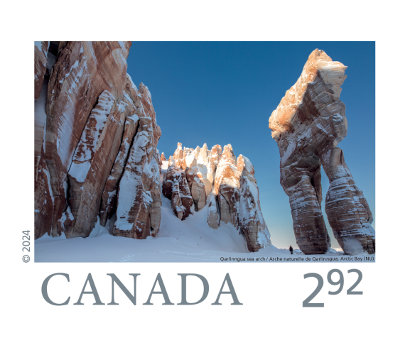 A stamp. Fantastic rock formations including fluted cliffs, but featuring a giant standing rock with two legs that looks like a pair of pants. A tiny figure stands beside it. It is winter and there is snow, and bright blue skies. The stamp reads “Canada 2.92” and “Qarlinngua sea arch/Arche naturally de Qarlinngua, Arctic Bay (NU)