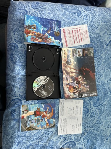 An open DVD case with a disc inside, alongside anime artwork covers and pamphlets placed on a blue patterned textile surface.