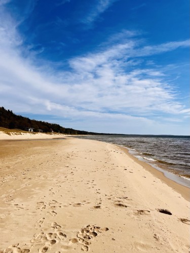 A sandy beach with footprints, bordering a calm Lake Michigan, under a blue sky with wispy clouds. Trees line the beach in the distance.
