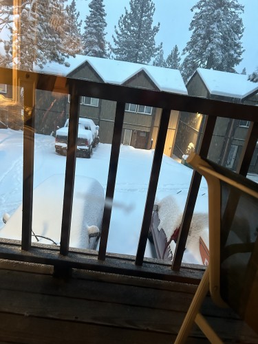 A view from a second story window. A porch deck of brown painted wood obscures the bottom half of the photo. A mesh fabric lawn chair is mostly out of frame on the right.

6 inches of snow fall covers a Lake Tahoe condo complex and its parking lot. Snow continues to fall. Snow covered pines tower 2 stories above the 2 story buildings. The two cars that face the window have their windshield wipers lifted to extend through the accumulating snow.

The light is dim blue outside. It is barely dawn. It is impossible to tell since the sky is heavy grey with clouds. It feels like the snow will never stop.