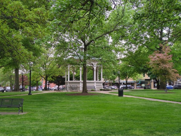 This photo shows a peaceful park setting featuring lush green grass, a variety of trees in full leaf, and a classic white gazebo in the center. The park is bordered by streets with parked cars and buildings in the background. Several pathways converge at the gazebo, and there are benches and lampposts scattered throughout the area.