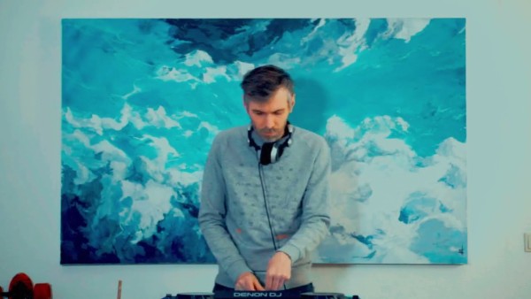 In the image, there is an individual who appears to be a DJ. They are focused on their work, with DJ equipment in front of them, including what seems to be a mixer or controller. They are wearing headphones around their neck and seem to be manipulating the controls of the device. The DJ is set against a large abstract painting or print with a motif resembling turbulent blue and white ocean waves or clouds, which occupies the wall in the background. The artwork gives a vibrant backdrop to the scene, suggesting a creative or energetic atmosphere. The setting seems to be indoors, possibly a home or studio space, and the individual is dressed in casual attire.