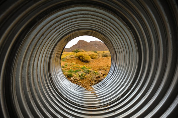 A photo of a metal spiral pipe with a desert background features where the pipe terminates, there are poppies and green shrubbery present with brown dirt and a small mountain range 