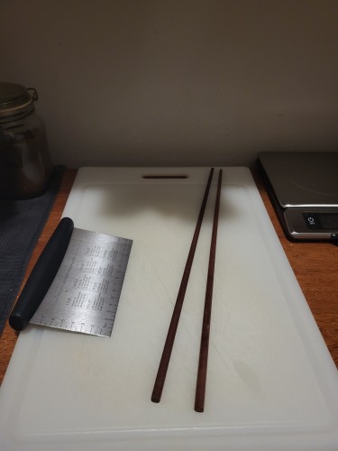Photograph of some cooking chopsticks on a cutting board, next to a bench scraper. They are almost as long as the cutting board is