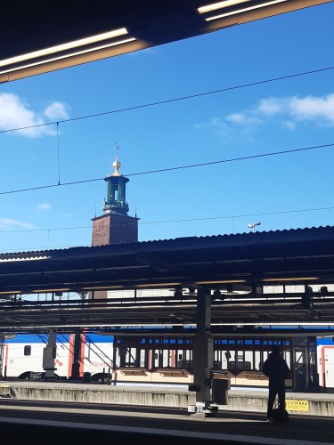 Photo of train tracks, with a train in the background. The sky is blue with a few small white clouds. In the background, a tower in red brick, a green bell cage on top, topped with a golden sculpture.