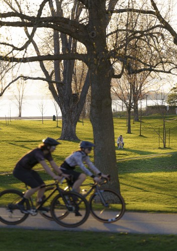 Two people on bikes ride by Britannia Beach in Ottawa, ON at sunset as a mother plays with her child in the background. Big tree dot the background with buds showing as it's spring 
