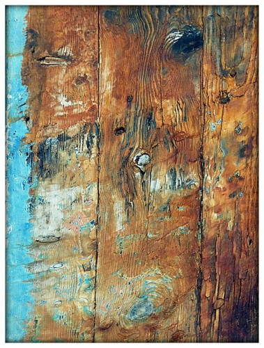 A colour photograph of old wood on a trawler which has stripped back from its blue covering, with only an edge of blue paint remaining in the left of the frame. The composition shows natural wood with various knots and remnants of paint in vertical sections transitioning to light blue paint.