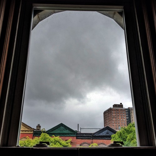 Looking through an arched window 150 minutes after sunrise the sky is full of gray storm clouds. Pointed roofs of Harlem brownstones with red brickwork are across the street, and a taller apartment building can be seen in the distance. The green tops of two trees are entering frame on the bottom and right.