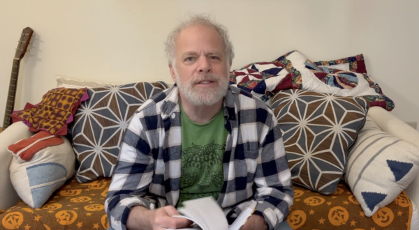 still from a video, showing a strikingly handsome White bearded middle-aged man sitting on a couch and flipping through a book