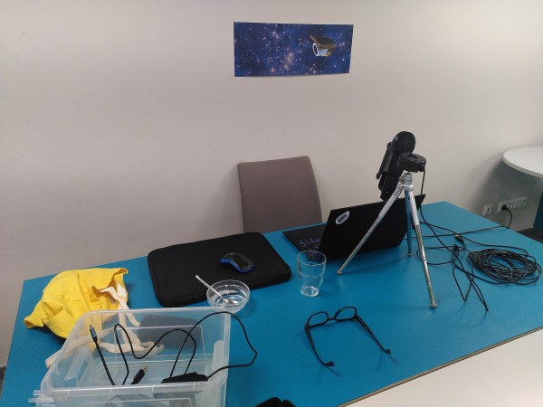 Desk with chair in front of white wall with a single picture. On the desk are a laptop, webcam on a tripod, microphone with pop-screen, as well as a glass of water, a pair of glasses and a box for equipment.