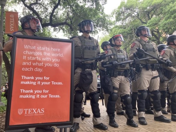 An echelon of police in riot gear stand with their batons at UT Austin behind a sign that states what starts at the university "changes the world" claiming that "it starts with you and what you do each day"
