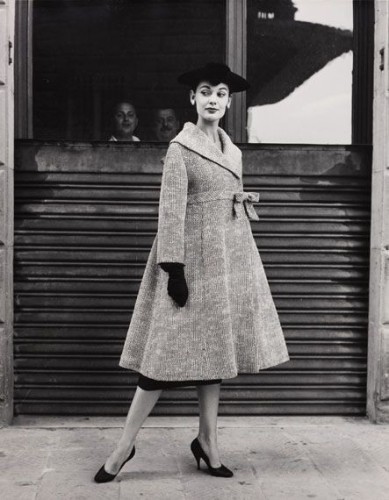  The image is a vintage photograph featuring a woman posing outdoors. She is dressed in elegant attire, including a coat with a fur collar and a hat adorned with a feather. Her posture is poised, with one leg slightly forward and the other bent behind her. The setting appears to be an urban environment, possibly near a building or storefront with large windows, as suggested by the reflection on the glass.

The woman's style and the overall composition of the photograph suggest that it could have been taken during the mid-20th century, capturing fashion trends from that era. The lighting is soft, indicating an overcast day or possibly early morning or late afternoon when shadows are longer. The image evokes a sense of history and the style of past eras, providing a glimpse into the fashion and lifestyle of the time.