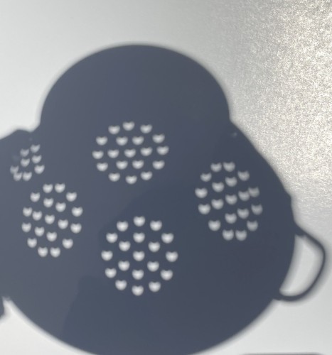 Shadow of a colander on white glossy paper. The colander is the kind with a stand on the bottom and round drainage holes pierced in circular groups. Each group of round holes, depending on its position in the colander, shows more or less of the bite taken by the moon out of the sun on the upward side of the image.