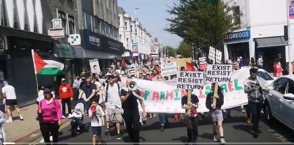 Huge crowd with flags, walking behind a banner reading STOP ARMING ISRAEL.  Two children at the front are holding placards reading EXIST RESIST RETURN