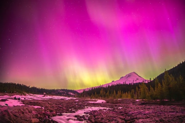 Pink and yellow light from the aurora borealis (northern lights) fills the sky above Mount Hood in this view from the White River West Sno-Park. The foreground shows a stream meandering through a rocky area.