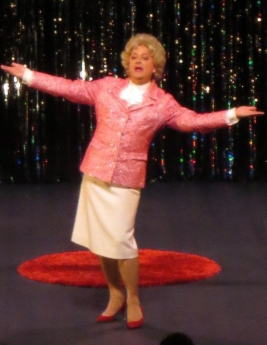 Photo de Marie-Thérèse Porchet les bras étendus, devant un fond scintillant, portant une veste rose brillante, une jupe beige et des chaussures rouges. Le fond est constitué de rideaux scintillants qui reflètent la lumière dans diverses couleurs. On aperçoit un tapis rouge circulaire au milieu de la scène.