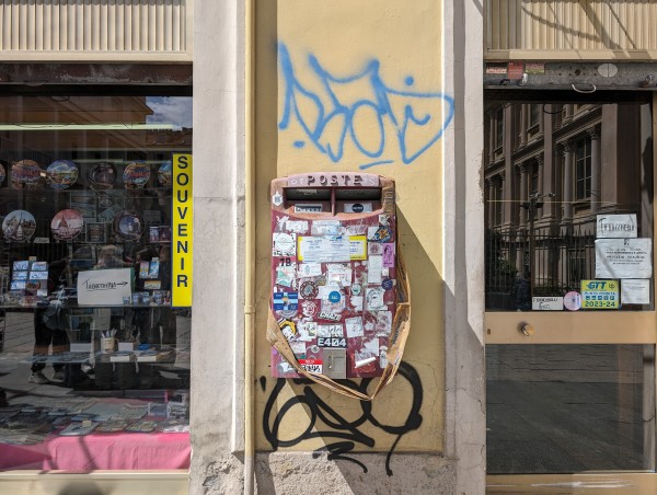 A much-stickered Italian mailbox mounted to a graffitied wall next to a souvenir shop. 
