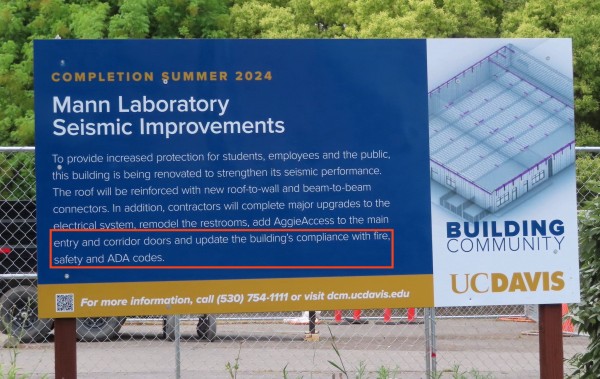 Large wooden sign board posted outside construction project. Features a bird's eye view of a floor plan. "Completion Summer 2024 Mann Laboratory  Seismic Improvements. To provide increased protection for students, employees and the public, this building is being renovated to strengthen its seismic performance.The roof will be reinforced with new roof-to-wall and beam-to-beam connectors. In addition, contractors will complete major upgrades to the electrical system. remodel the restrooms, add AggieAccess to the main entry and corridor doors and update the building’s compliance with fire, safety and ADA codes. For more information, call (530) 754-1111 or visit dcm.ucdavis.edu" "Building Community UC Davis"