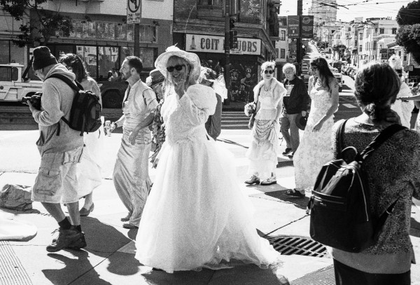 a scan of a black and white film negative of women dressed in bridal dresses, waving and walking around san francisco