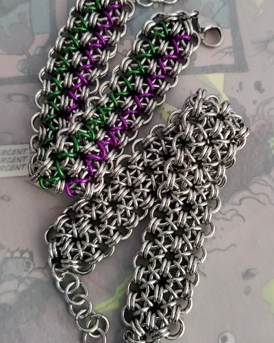 two chainmail bracelets folded in half laying next to each other. they are designed with a lace weave and are wide like a cuff. upper one is stainless steel paired with purple and green anodized aluminum.