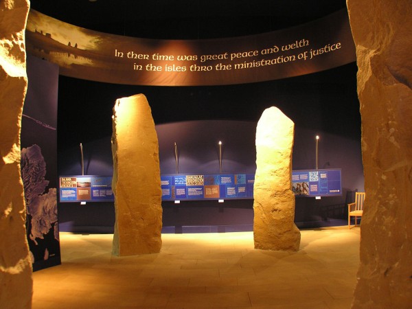 The Museum of the Isles at Armadale Castle. The image shows a room with dark walls and ceiling and interpretive boards along the back wall at waist level. The main body of the image is filled with four large standing stones. Two are seen fully while the other two form the edges of the image. A curved panel below the ceiling talks of the Lords of the Isles.