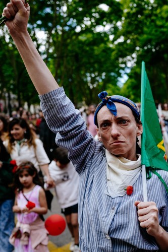 A person raising their fist in a defiant gesture, wearing a striped shirt with a red flower pinned to it and a blue headscarf, holding a green flag. Background shows a crowd with some children and trees.