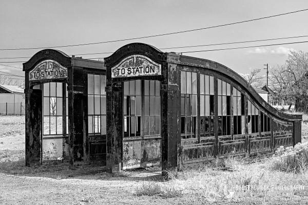 Black and white photo of two old art-deco style entrance canopies for an underground metro system. They sit side-by-side in a vacant lot with a few weeds. About half of the windows are broken. Over the top of each canopy is says, "To Station." 