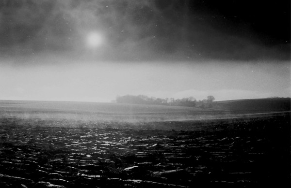 Photo en noir et blanc d'un champ fraîchement labouré une brume d'élève du sol, la soleil commence à apparaître au travers de la brume