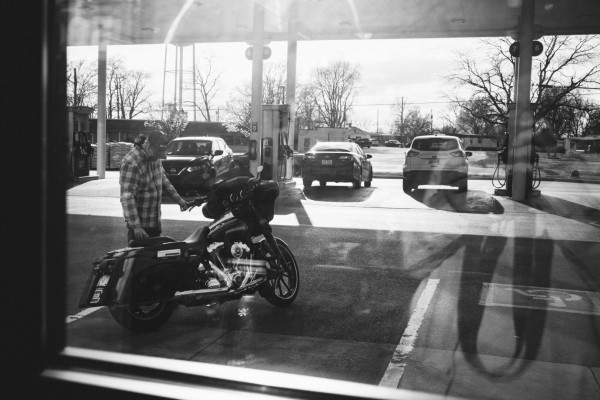 The photo is a black and white image capturing a moment at a gas station. The main subject is a person standing next to their motorcycle parked in a space in front of the gas station, their face obscured by a baseball hat. The photo is taken from inside the station through a window. The scene is busy with several cars in the background, and the strong sunlight light creates silhouettes of the cars outside and reflections of the photographer in the glass, adding depth to the image. The gritty monochrome effect gives the image a timeless, nostalgic feel.