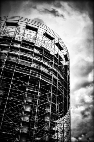 Looking up at a large water tower under repairs, it is surrounded with scaffolding in this black and white photo.