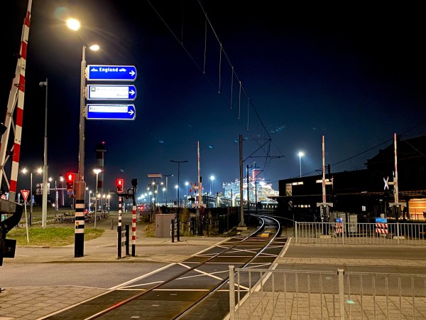 Exterior, night. A railway track runs through the center of the scene, fences and barriers protecting a street crossing it in the foreground. A sign poins to the boat to England. In the distance a large, illuminated ship can be seen.