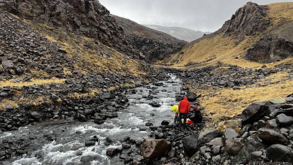Three people in red, yellow, and black jackets by a rocky stream in a mountainous area. The sky is overcast. 