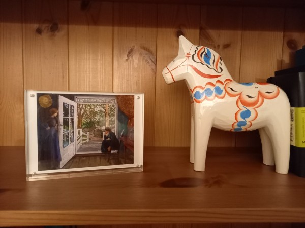 Ausschnitt eines Bücherregals. Links steht ein Bild (Nikolai Astrup "Bei der offenen Tür") in Postkartenformat im Plexiglasständer, rechts ein rotblauweißes Dala-Pferd.