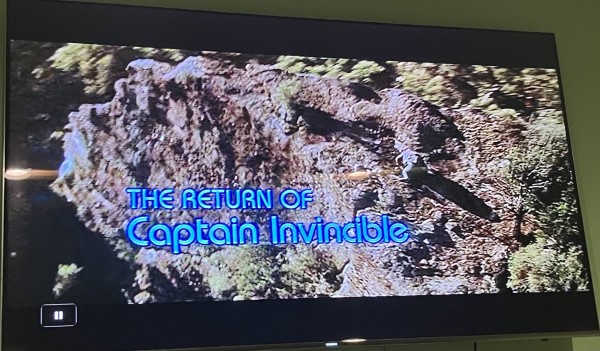Title card for The Return of Captain Invincible. Alan Arkin or his stunt double has just fallen over backwards & is struggling to rise again, butt first. 