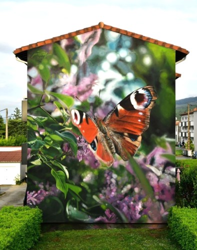 Streetartwall. A fantastic, photorealistic mural with a butterfly in a green landscape was painted on the exterior wall of a modern two-storey residential complex. A large brown and white butterfly sits on small green leaves. The mural is designed like a photograph, with the surroundings behind the butterfly slightly blurred. Purple flowers (lilacs) and green foliage can be seen there.
Info: The Artist "Mantra" aka Youri Cancell, using rollers and brushes to apply acrylic painting to the multistory residential building. 
Butterfly Name: Peacock butterfly
Binomial name: Aglais io
Flowers: Syringa vulgaris