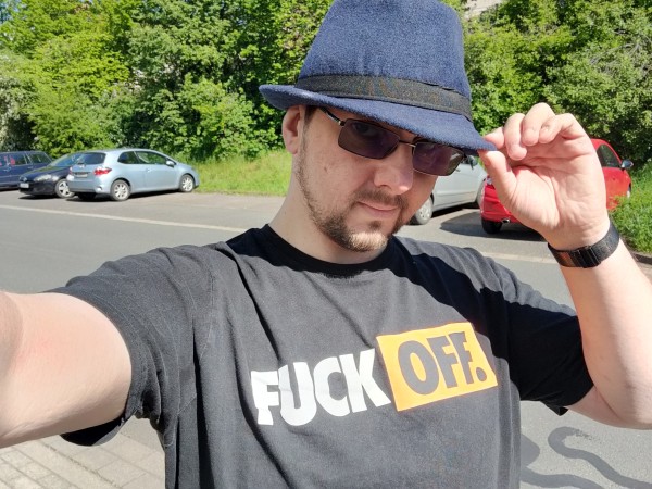 Ich halte meinen blauen Hut mit der einen Hand fest und mache ein Selfie. Ich trage ein schwarzes T-Shirt mit der Aufschrift Fuck off