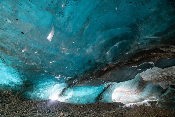 An interior view of a glacier cave with a stunning blue light emanating from two entrances on either side,
 illuminating the ice walls and layers of rocky inclusions through the crystal clear blue ice. 