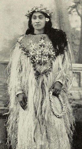 This is a photo from 1906 of a beautiful Tahitian woman.

The image is in black and white. It shows a woman in her twenties standing tall. Her body is slightly tilted to the right, her hands resting on her hips. In her left hand is a wreath made of flowers.

Her outfit is a long gown seemingly made of some kind of plant fibre. It is finely woven in a fishing net pattern on her chest and decorated there with bows and flowers made of plant fibre.

The garment is loose and feels tasselled.

Her dark, thick hair is unbound and falls over her shoulder to chest length.
She wears a flower crown.

Her face is pleasant, with a slight smile and large dark eyes, with well-shaped eyebrows.

A single lotus flower adorns her right ear.