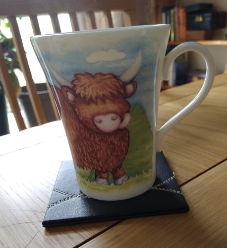 Mug of tea on a table, with a highland cow licking it's lips! The is blue sky behind it with a white cloud.  It is standing in green grass 