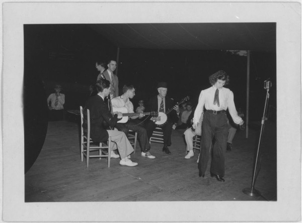  This black and white photograph captures a lively scene of musicians performing at the Mountain Music Festival in Asheville, North Carolina. A group of people is immersed in their performance on stage. The musicians are actively engaged with their instruments; some are playing guitars while others are using banjos or fiddles. The audience members in the foreground seem to be enjoying the live music. The image exudes a sense of nostalgia and captures a moment in time when music brought people together in celebration.

The photograph, dated between 1950-1970, serves as a testament to the rich musical heritage of Asheville. It's a snapshot of a bygone era, where the joy of music transcended age, culture, and geography. The image is a window into the past, offering a glimpse of the vibrant cultural life that thrived in the Blue Ridge Mountains of North Carolina during that period. 
